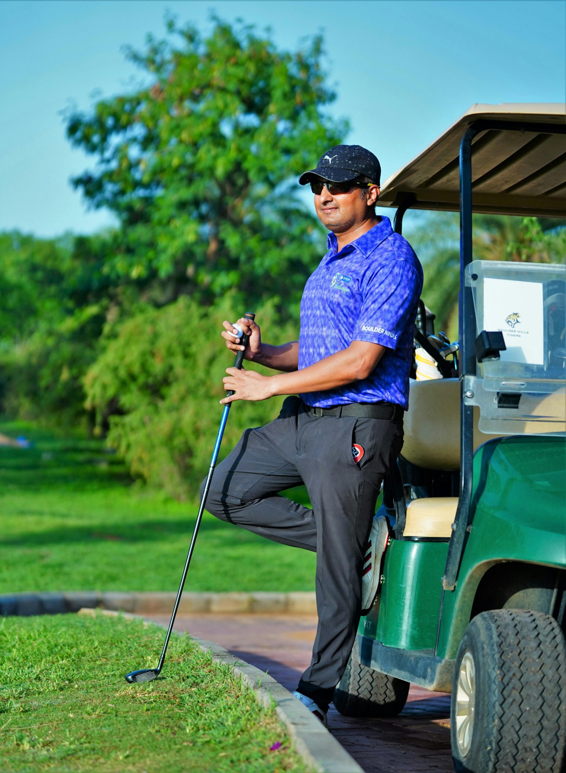 A man in sportswear stands with a golf club beside a cart on a lush golf course.