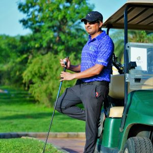 A man in sportswear stands with a golf club beside a cart on a lush golf course.